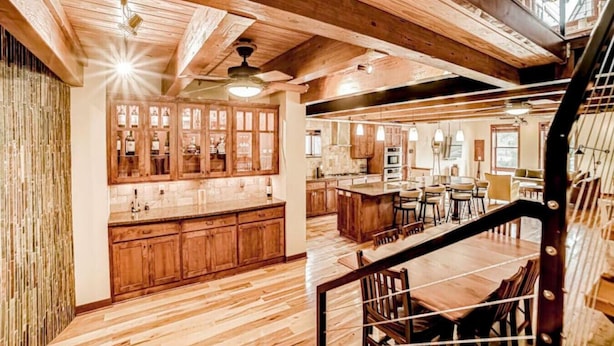 Un'ampia cucina con mobili in legno e un'isola centrale, un'area pranzo con tavolo e sedie in legno, e una scala che conduce a un piano superiore.