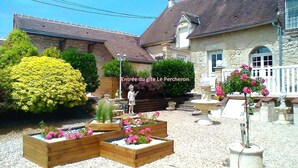 A garden with wooden planters, a statue, and a stone building with a tiled roof. - Gîte in the heart of the Perche, Le Percheron (Perche-en-Nocé)