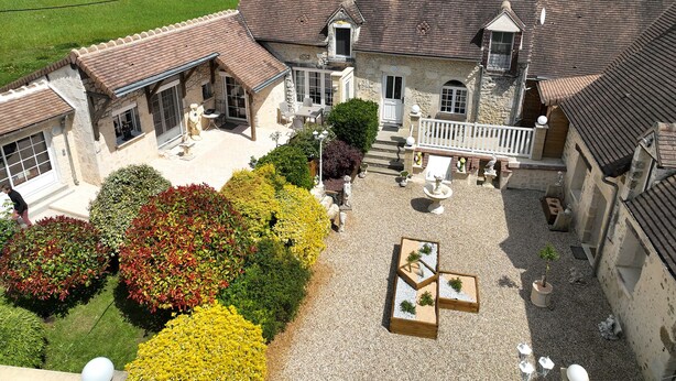 A large stone house with a courtyard, featuring a dining area, outdoor seating, and a garden with various plants. - Gîte in the heart of the Perche, Le Percheron (Perche-en-Nocé)