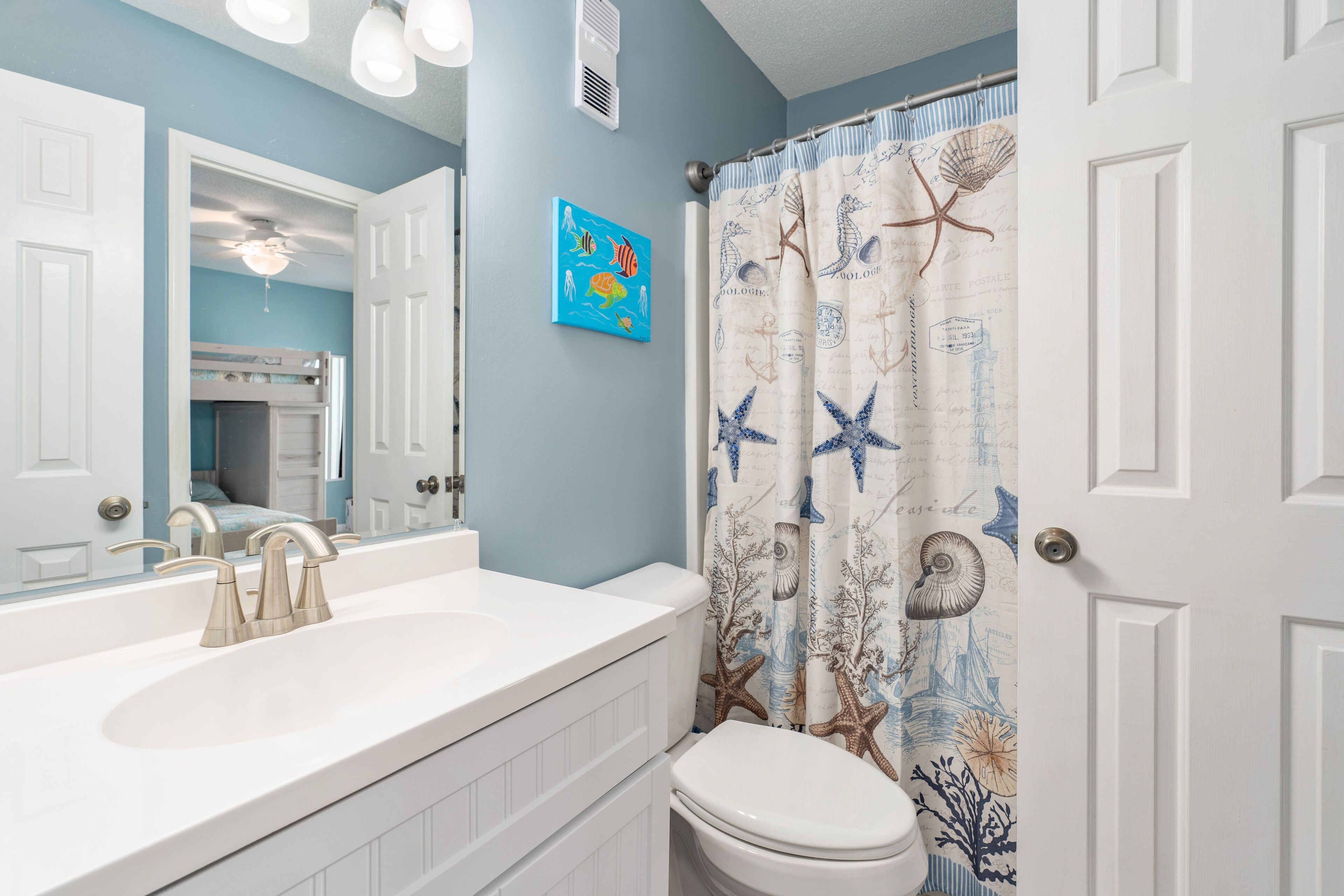 Een badkamer met een wit wastafel, een spiegel en een toilet. De muur is blauw en er is een douchegordijn met een nautisch thema.