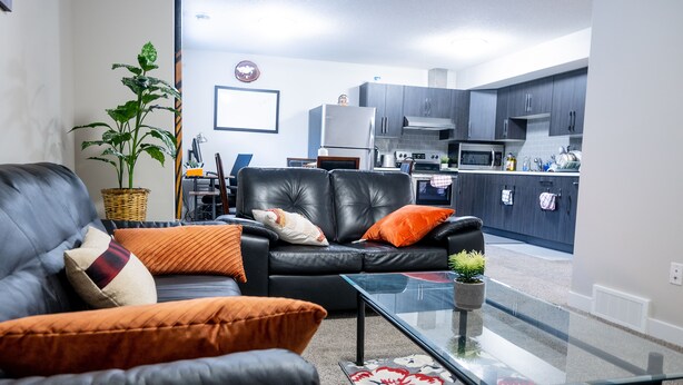 A living room with a black leather sofa, orange cushions, a glass coffee table, and a potted plant. - Exquisite & Cozy La'Posh Suite w/WiFi, Workspace, Storage (Airdrie)
