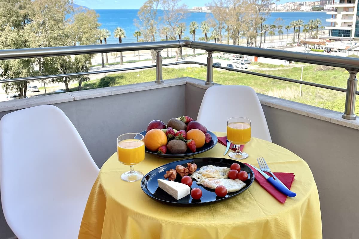 A balcony table with breakfast items, a view of the sea, and a building in the background.