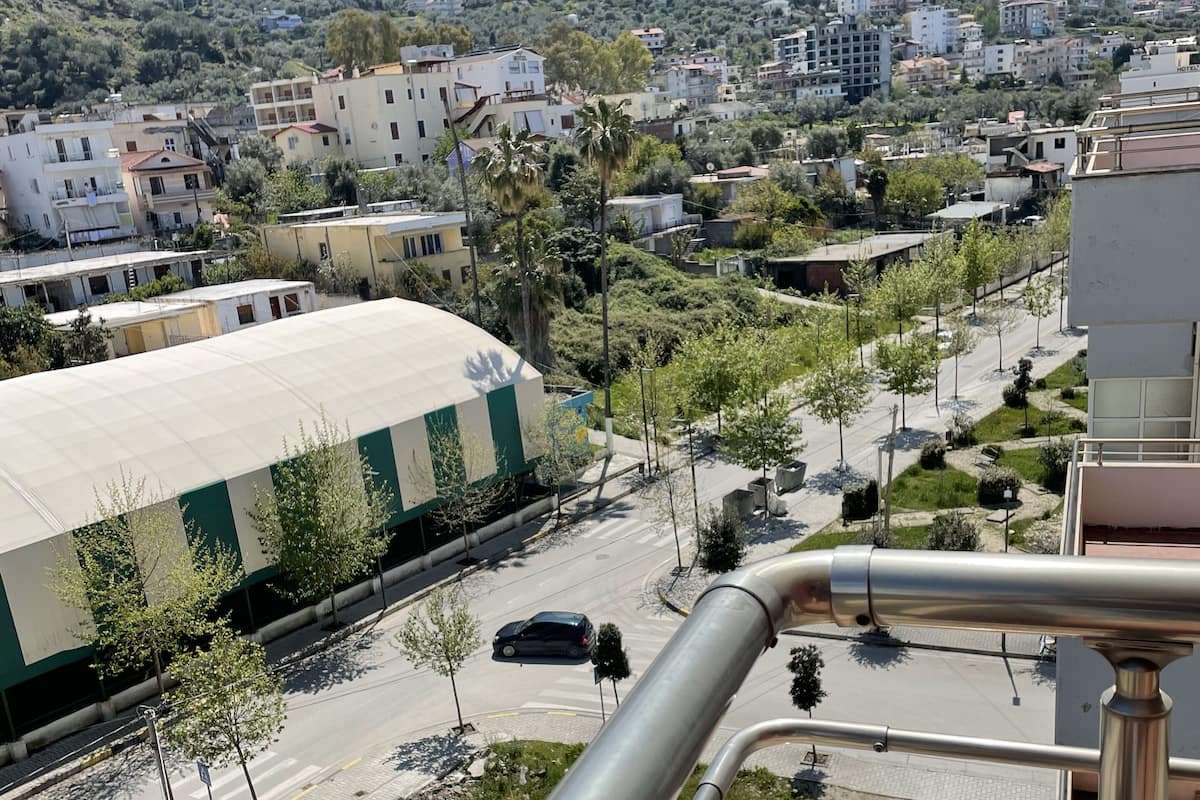 A view from a balcony overlooking a street with buildings, trees, and a mountain in the background.