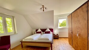 A bedroom with a wooden floor, a bed with white linens, a red headboard, and a window with a view of trees.