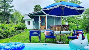 A pool area with a blue inflatable raft, a shark float, and a blue umbrella. There is a house in the background with a porch and outdoor furniture.