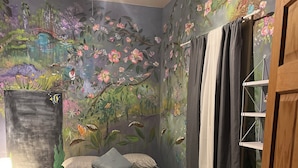 Una camera da letto con un letto, un comodino, una lampada, una finestra con tende e un affresco.