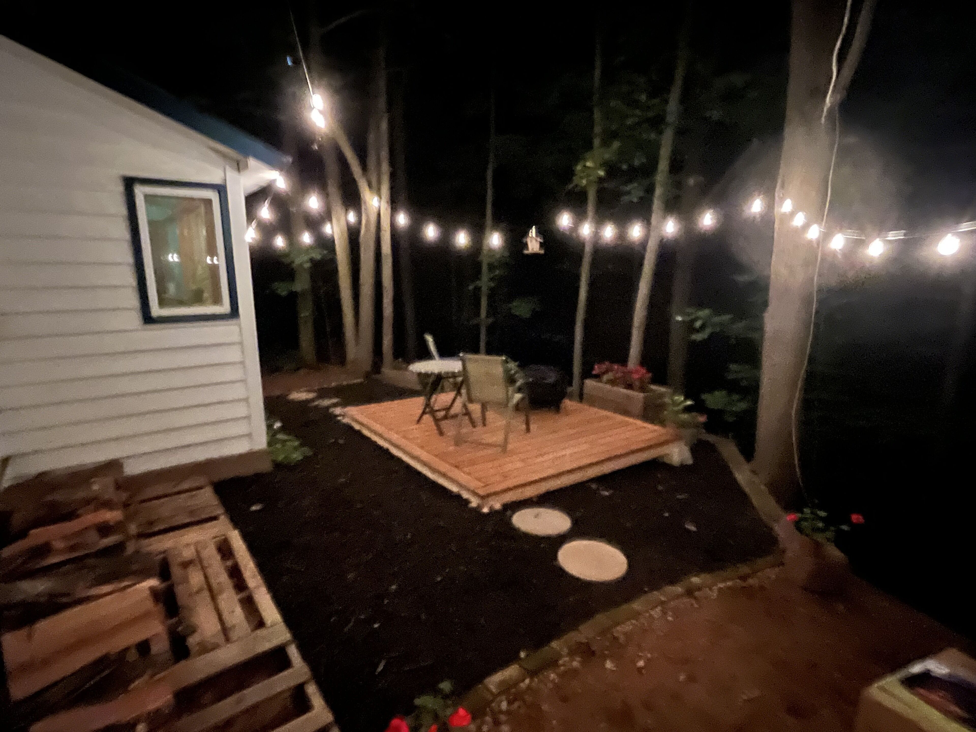 Un patio trasero con terraza de madera, una mesa y sillas, y luces colgantes.