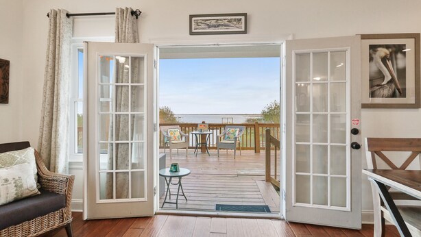 A room with a view of a wooden deck and a balcony. There is a wicker chair, a small table, and a framed picture on the wall.