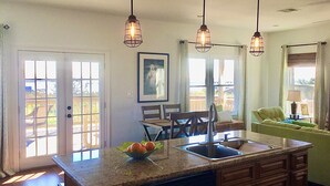 A kitchen with a central island, a dining area with chairs, and a view of the outdoors through large windows.