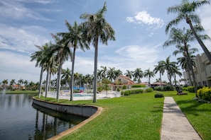 A landscaped area with a pond, palm trees, a pathway, and a building in the background.