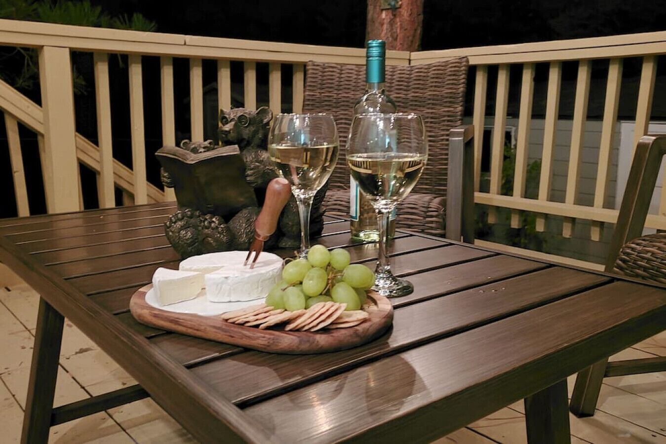 A wooden table set with a cheese wheel, grapes, crackers, and two wine glasses with a bottle of white wine.