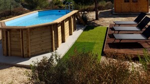 A wooden swimming pool with a wooden deck, surrounded by a grassy area and a small wooden cabin.