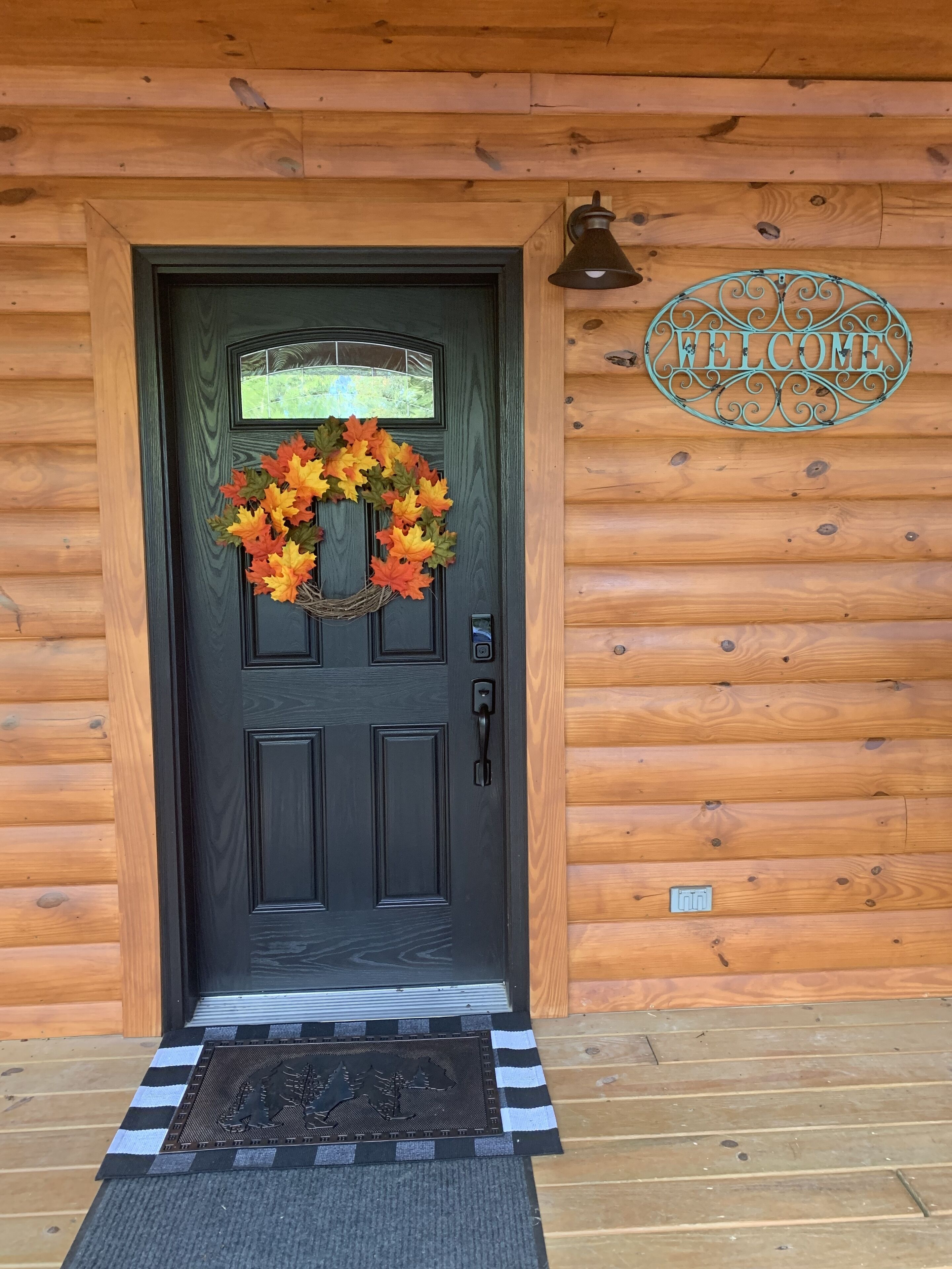 A wooden cabin door with a black door, a welcome sign, a wreath, and a doormat.