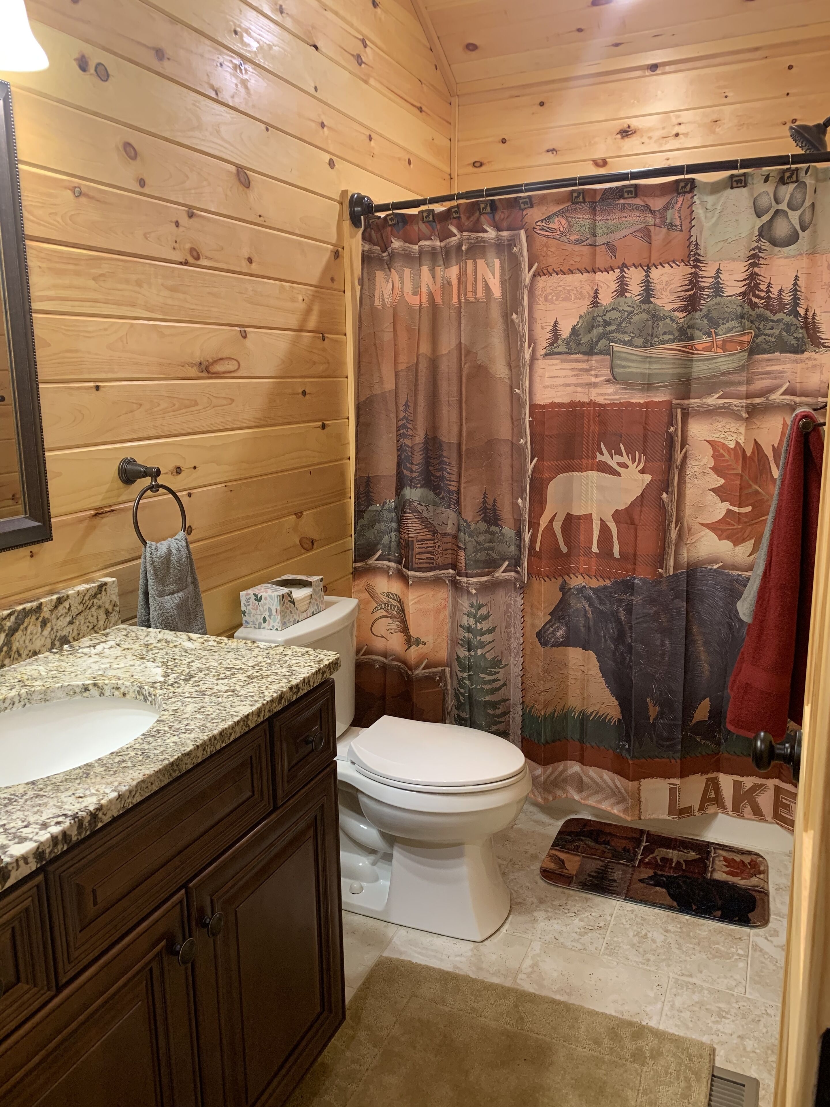 Een badkamer met houten wanden, een granieten aanrecht, een toilet en een douchegordijn met een ontwerp van een bos en dieren.