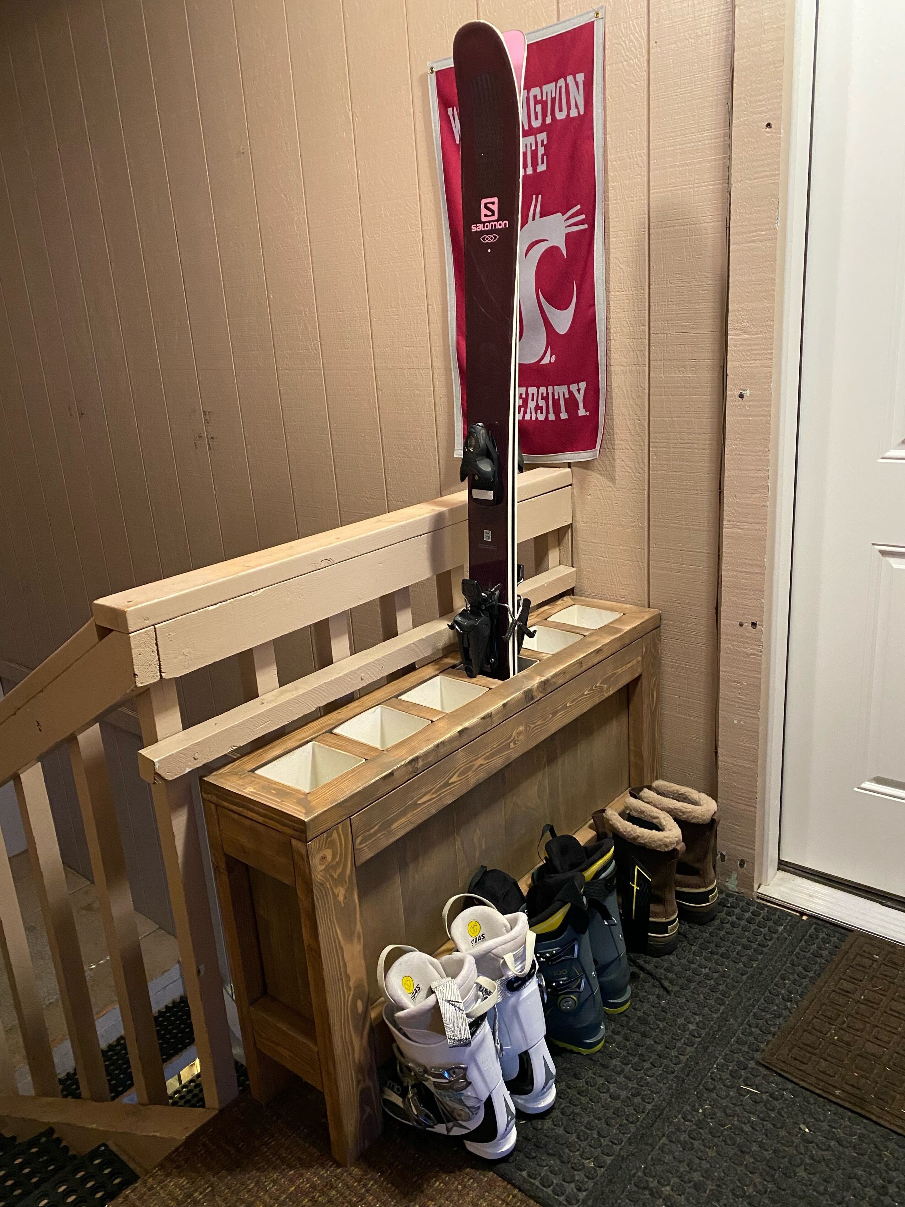Een houten bankje met een snowboard tegen een muur, een deur en een schoenenrek met laarzen.