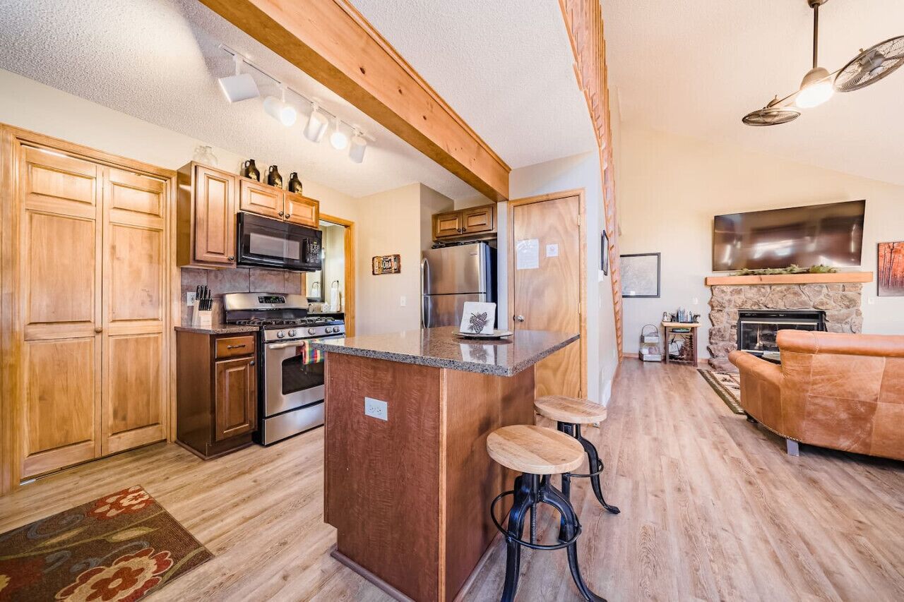 A spacious kitchen with wooden cabinets, a central island with stools, a built-in oven, and a stone fireplace.. Walk to National Park! Mountain Cabin w/ Fireplace