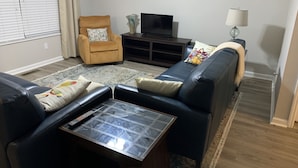 Un salón con sofá, sillón, mueble para televisión y una mesa de centro de vidrio.