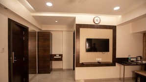 A modern hotel lobby with a flat-screen TV mounted on the wall, a clock, a reception desk, and a bench. - Hotel Naaz Executive Near T2 Airport (Mumbai)