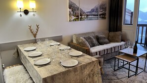 A dining area with a wooden table set for six, a sofa with cushions, a small table with glasses and a bottle, and a painting of a snowy landscape on the wall.