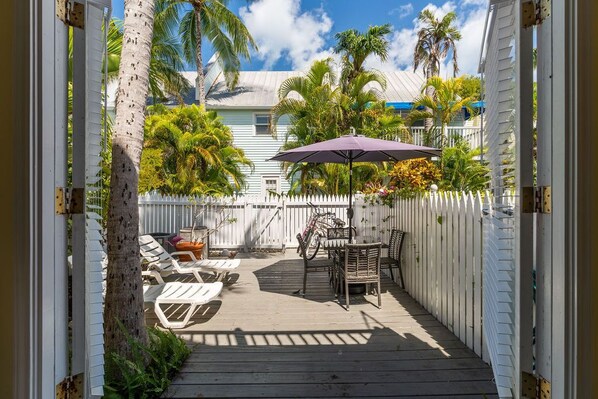A wooden deck with lounge chairs and a table, surrounded by a white picket fence and palm trees.