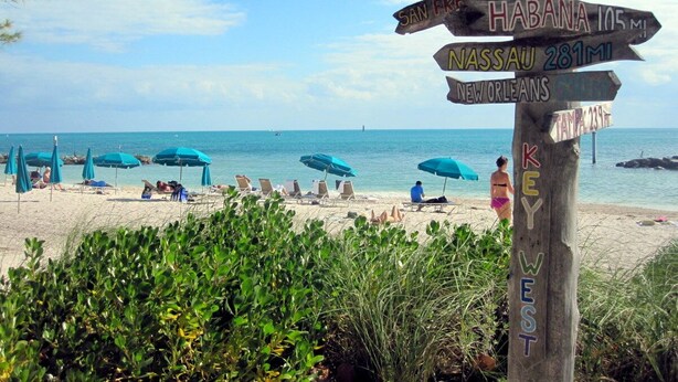 A beach with lounge chairs and umbrellas, and a wooden signpost with city names and distances.