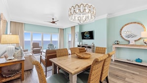 A spacious living room with a wooden dining table, wicker chairs, a large window with a view, a ceiling fan, and a mounted television.