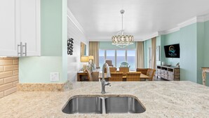 A modern kitchen with a granite countertop and stainless steel sink, a dining area with wicker furniture, and a view of the ocean through large windows.
