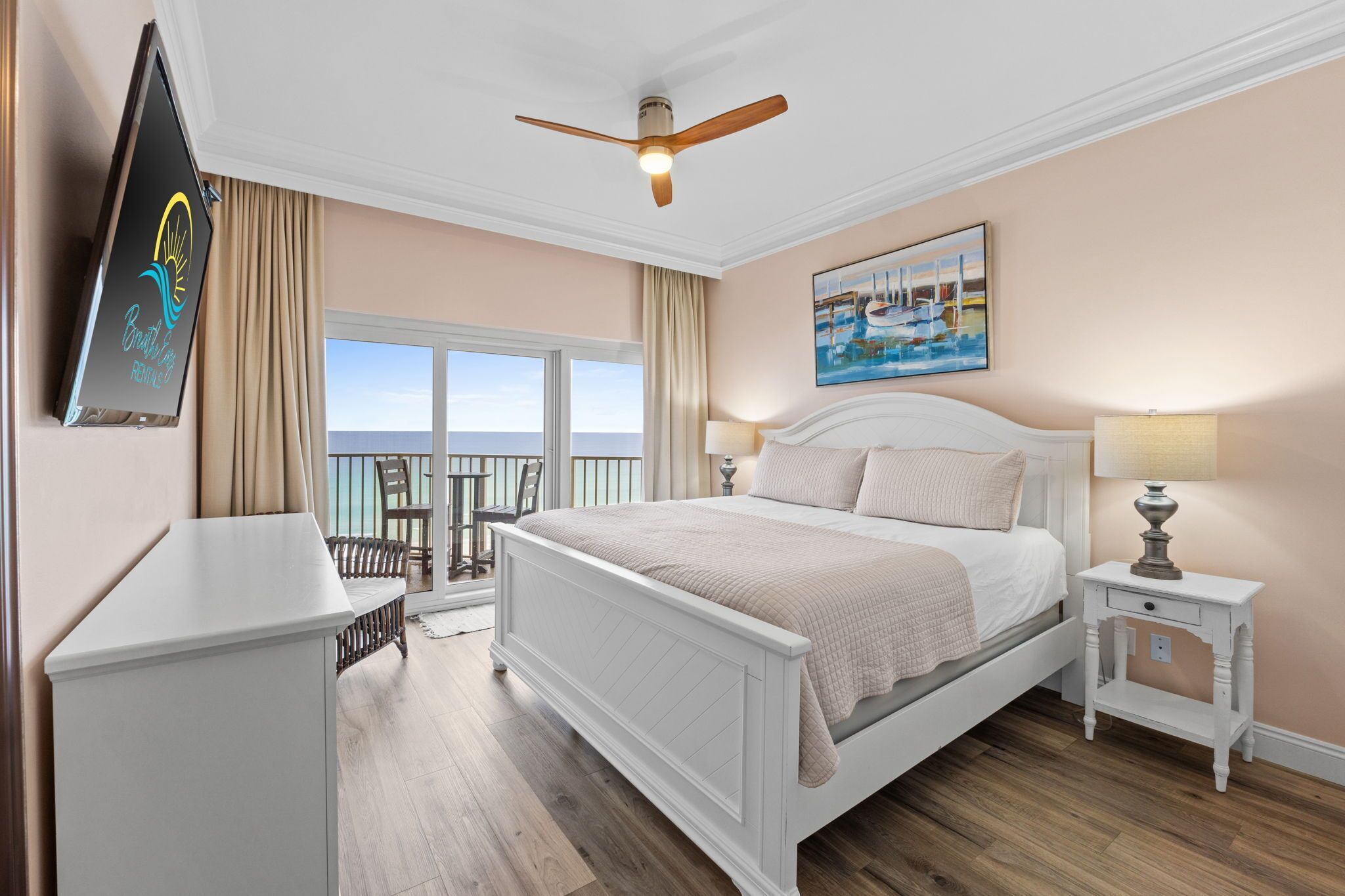 A bedroom with a bed, a nightstand, a desk, a chair, a ceiling fan, a painting, and a view of the ocean.
