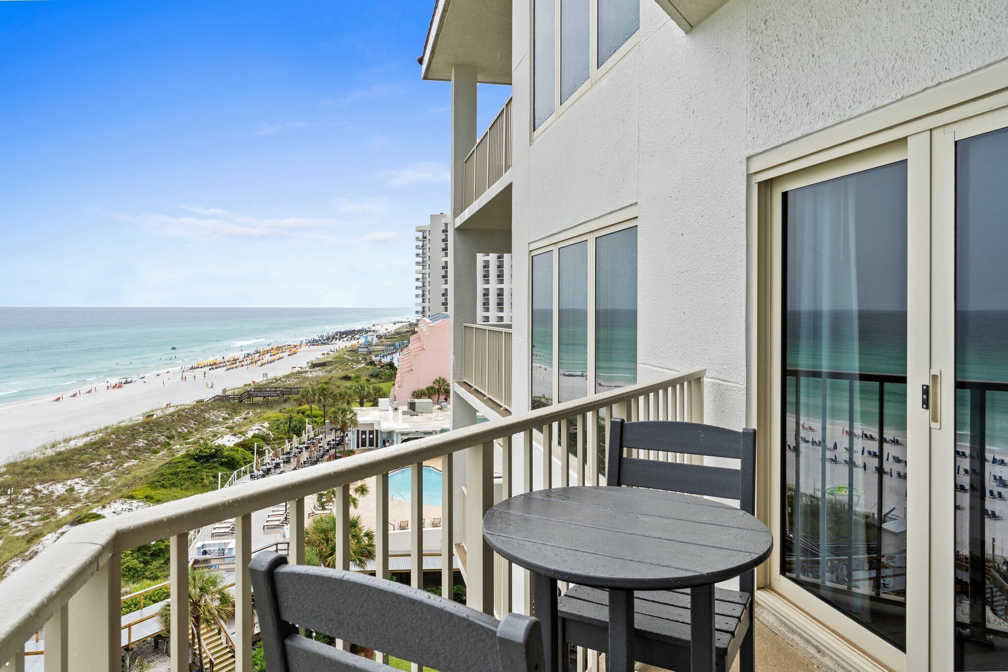 A balcony with a table and chairs overlooking a beach with a pool and buildings in the background.