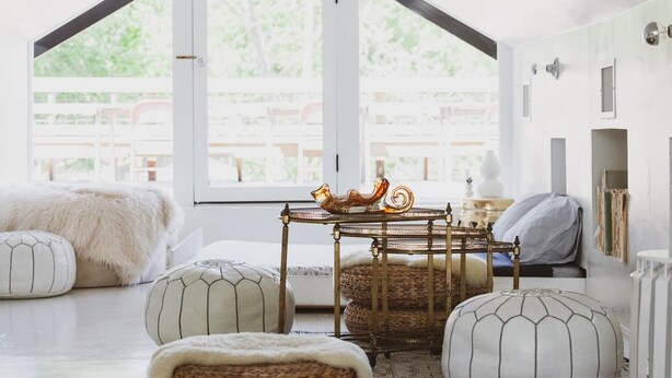 A cozy room with a high ceiling, a large window, and a view of outdoor furniture. There are two white upholstered chairs, a small table with a decorative item, and a fluffy white rug. - Spacious and Charming- Walk to Rainey Street (Austin)