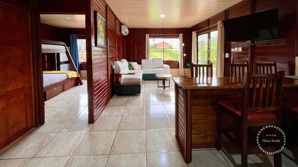 A spacious living area with a sofa, a dining table, and a TV. There is a staircase leading to a bunk bed area. - Cozy villa surrounded by nature with AC & pool (La Fortuna)