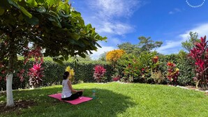 A person meditating on a pink mat in a garden with various plants and a clear blue sky. - Cozy villa surrounded by nature with AC & pool (La Fortuna)