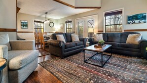 A spacious living room with a dark leather sofa, a grey armchair, and a coffee table. There are large windows with blinds, a dining area in the background, and a chandelier.
