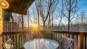 A wooden deck with a glass table and chairs, overlooking a forested area.