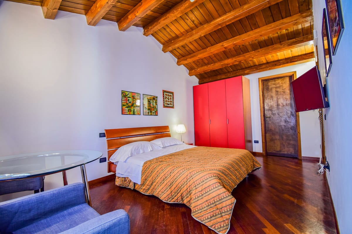 A bedroom with a wooden ceiling, a glass table, a bed with a patterned blanket, a red wardrobe, and two wall-mounted artworks.