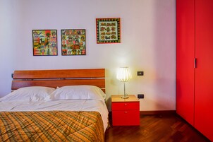 Un dormitorio con una cama de madera, una mesita de noche roja con una lámpara y dos cuadros enmarcados en la pared.