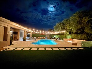 Un patio trasero con piscina, muebles de exterior, luces colgantes y vistas al cielo nocturno.