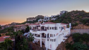 A large, multi-story white building with balconies, situated on a hillside with other buildings in the background.
