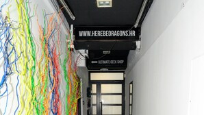 Un corridoio con pavimento in legno scuro, un'installazione artistica astratta e colorata sulla parete sinistra, e un cartello sopra la porta che recita 'ULIMATE GEEK SHOP'.