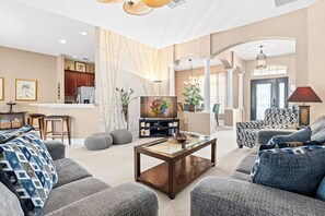 A spacious living room with a large sofa set, a glass coffee table, and a television. There is a kitchen area in the background with wooden cabinets and a refrigerator. - Minutes to Disney World & Margaritaville -WiFi-Game Room-Private Saltwater Pool  (Kissimmee)