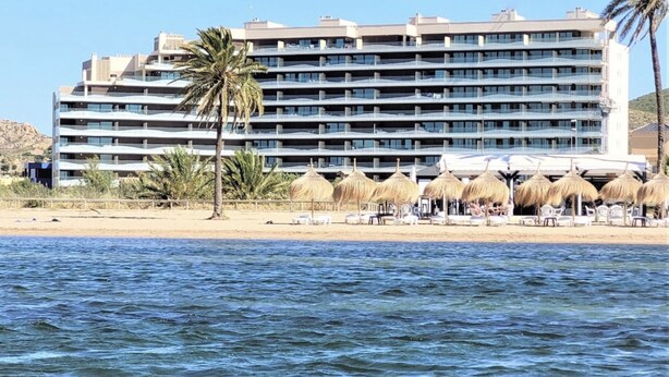 Een hotel met meerdere balkons, een strand met rieten parasols en een palmboom.