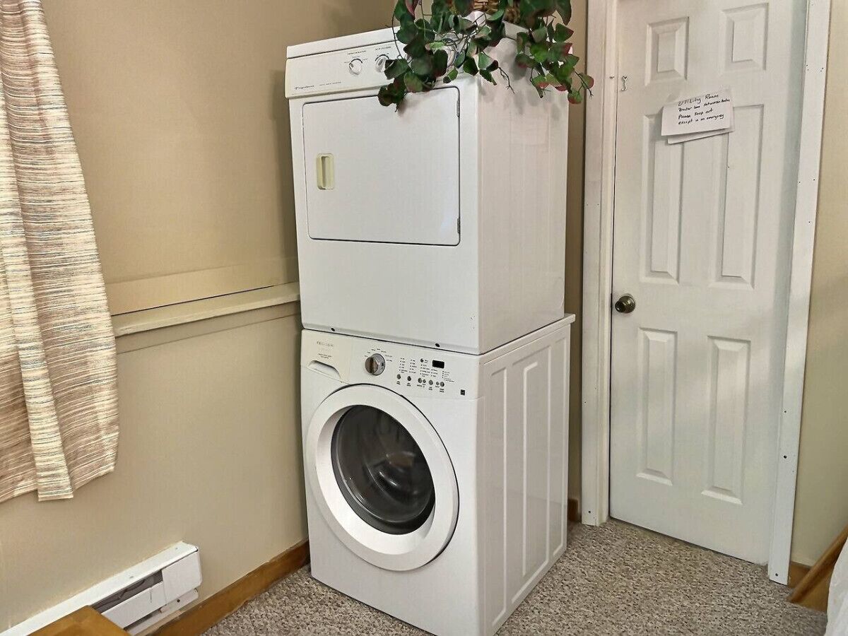 Un appareil combiné lave-linge et sèche-linge, installé en hauteur, se trouve dans un couloir, avec une porte sur le côté droit.