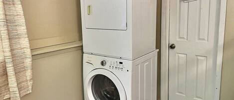 Un appareil combiné lave-linge et sèche-linge, installé en hauteur, se trouve dans un couloir, avec une porte sur le côté droit.