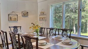 Un comedor con una mesa redonda dispuesta para seis personas, rodeada de sillas ornamentadas. Hay cuadros en la pared, un florero con flores sobre la mesa y vistas a árboles a través de amplias ventanas.