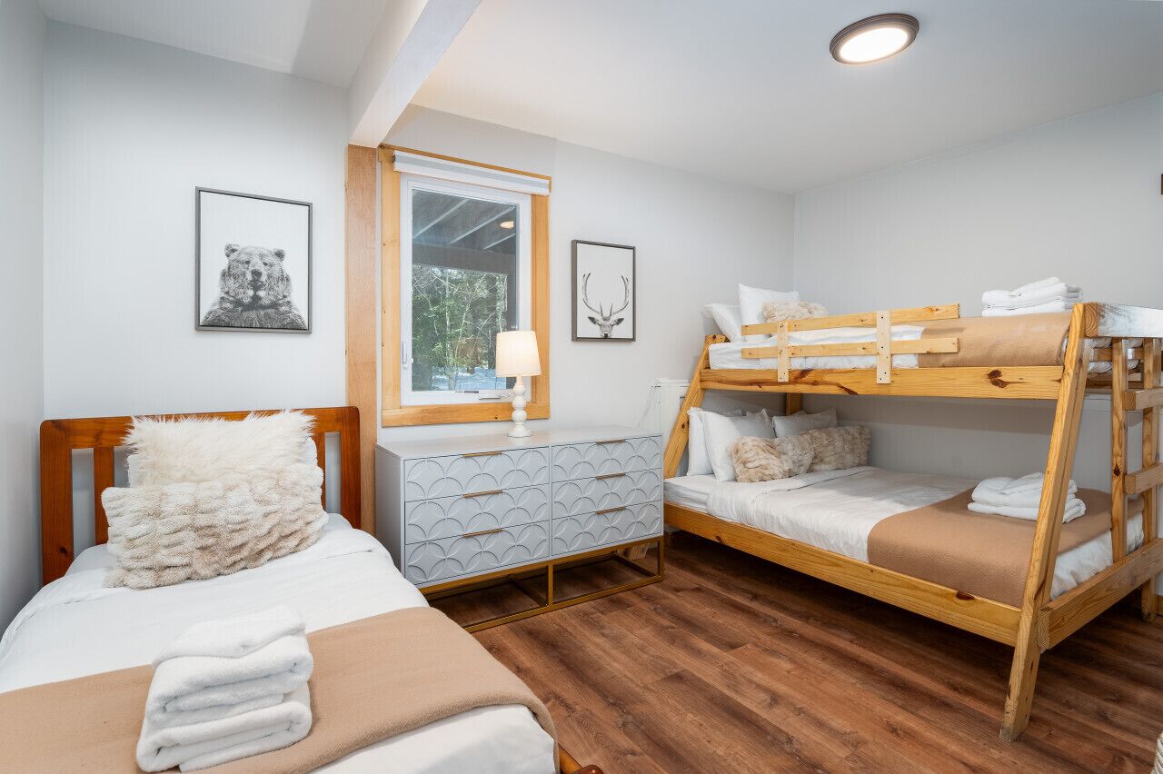 A room with two beds, one with a wooden headboard and the other with a bunk bed. There is a dresser with a patterned drawer front and a window with a view of greenery outside.