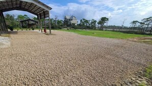A gravel-covered area with a covered structure, a grassy field, and a house in the background. - Waterfront RV Site with Dock (Port St. Joe)