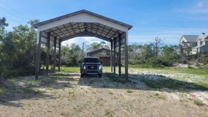 A covered outdoor area with a car parked underneath, surrounded by natural vegetation and a residential building in the background. - Waterfront RV Site with Dock (Port St. Joe)