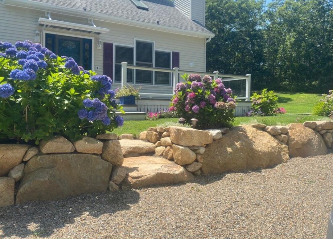 Une maison avec un mur de soutènement en pierre, une allée en gravier et un porche à l’avant, agrémenté de plantes en pot.