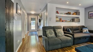 Una sala de estar moderna con un sofá gris, suelo de madera y una estantería empotrada con libros y adornos.