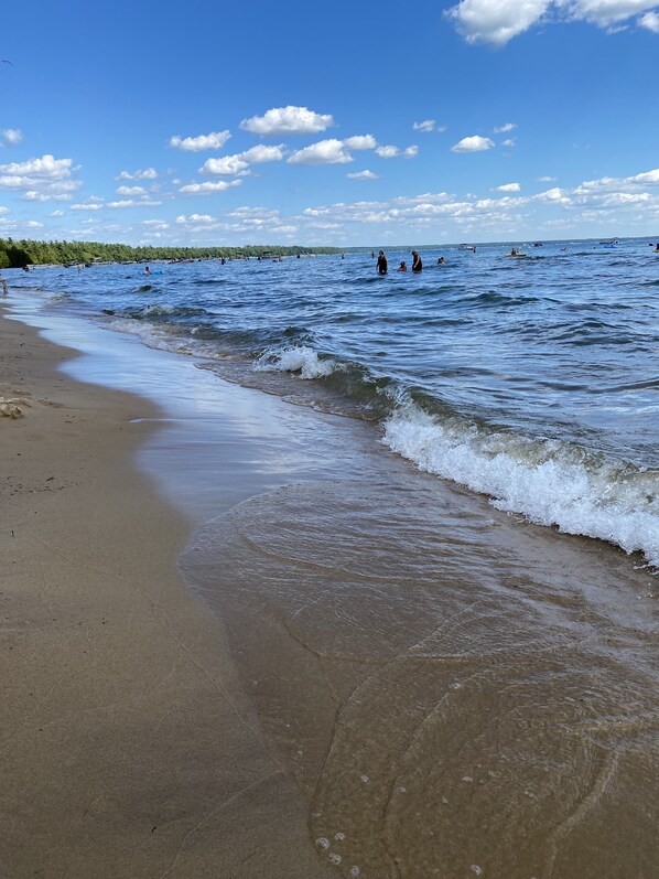 Uma praia com ondas suaves quebrando na costa, céu claro e pessoas nadando à distância.